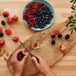 Overhead shot of fresh fruits, nuts, and hands preparing ingredients on a wooden board.